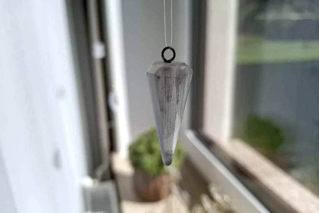 Pale faceted pendulum hanging from a thin line in front of a window.