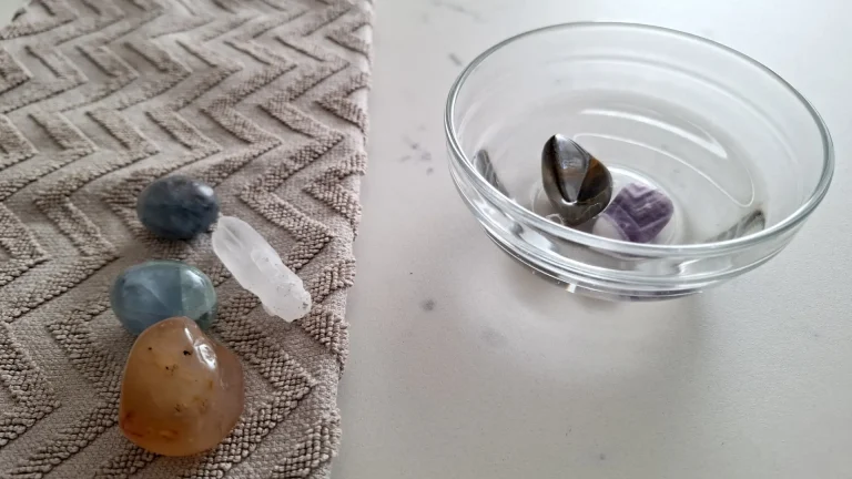 Tumbled stones and a clear quartz point on a towel beside a glass bowl of stones in water.