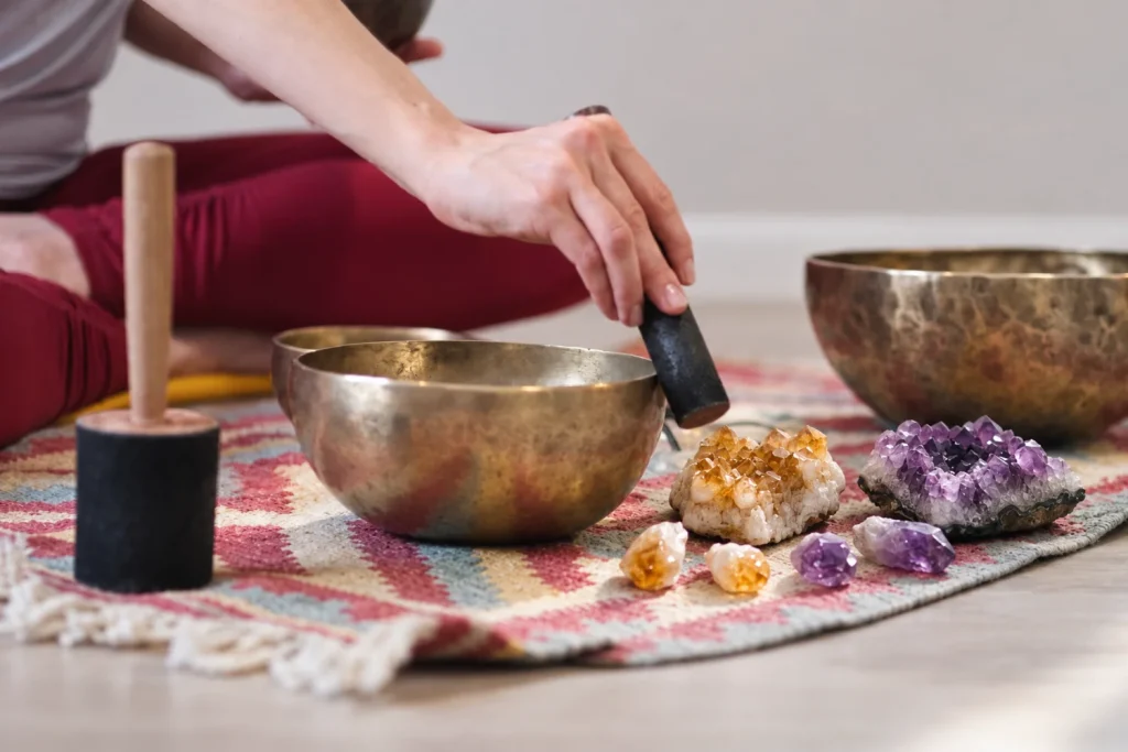 Person seated on the floor striking a brass singing bowl, with citrine and amethyst crystals on a woven rug