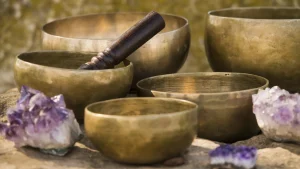 Five brass singing bowls on a stone ledge with a wooden mallet and amethyst clusters beside them