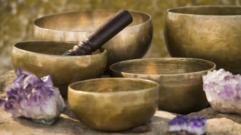 Five brass singing bowls on a stone ledge with a wooden mallet and amethyst clusters beside them